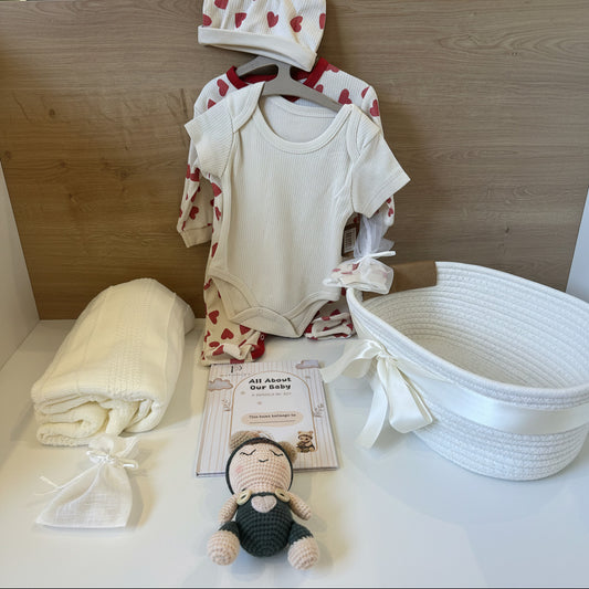 A baby gift set arranged on a white surface against a wooden backdrop, including a cream ribbed bodysuit with red heart print on a hanger, a matching bonnet, a folded cream blanket, a small plush doll, a card reading 'All About Our Baby,' and a white woven basket.