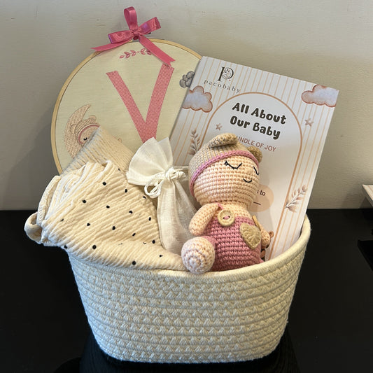 A baby gift basket set containing a cream crocheted basket, a pink and white crochet teddy bear, a book titled 'All About Our Baby', and a round embroidered patch with a pink bow, arranged on a dark surface against a light wall.