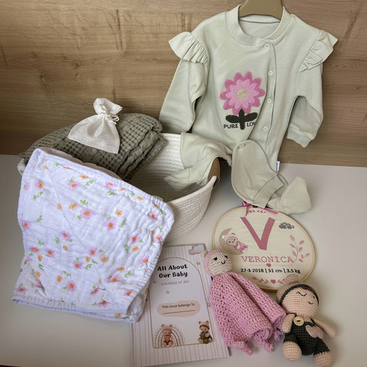 A baby gift set arranged on a light surface against a wood-paneled background, including a pale sage green baby romper with a pink flower graphic, a floral muslin blanket, a folded gray knit item with a small white bow, a round embroidered name piece reading 'VERONICA', and a pink crocheted toy.