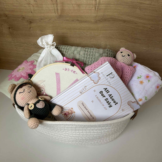 A baby gift basket arranged in a white woven basket, containing a crocheted monkey doll, a pink plush bear, a floral baby blanket, a knitted gray wrap, a small white drawstring bag, and printed cards. The basket is set against a light wood-paneled background.
