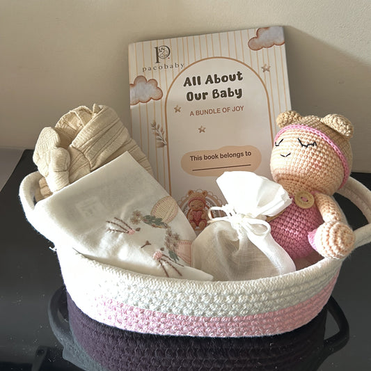 A baby gift set arranged in a crocheted basket on a shelf, including a knitted bunny plush toy, a book titled 'All Hearted Baby', and a swaddled baby blanket.