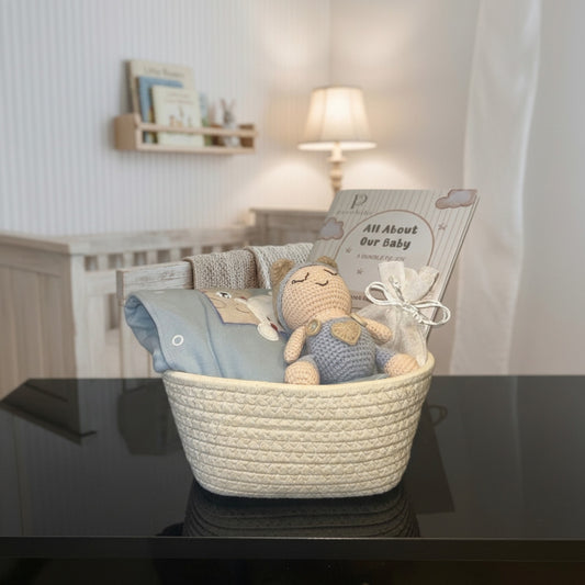 A baby gift basket set in a nursery, featuring a cream woven basket containing a blue knit blanket, a crocheted teddy bear, and a gift bag with a card that reads 'All About Our Baby'.