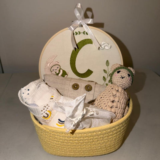 A baby gift basket set featuring a yellow woven basket, a round wooden monogrammed lid with a green letter C, a beige linen pouch, a knitted beige bear toy, and white baby clothes inside.
