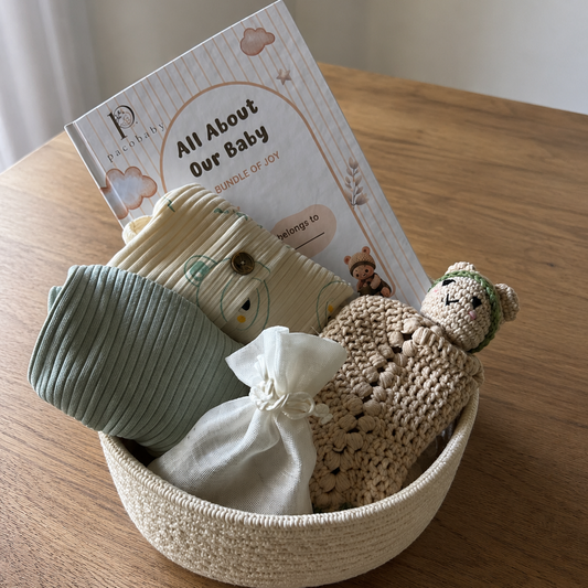 A baby gift set arranged in a woven cream basket on a wooden table, featuring a sage green ribbed swaddle, a beige plush toy, a small white drawstring pouch, and a printed booklet titled 'All About Our Baby'.