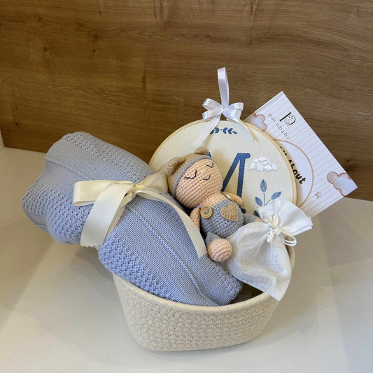 A baby gift basket containing a light blue knit blanket tied with a cream ribbon, a small plush toy, a round embroidered patch, and a printed card, arranged in a cream woven basket against a wooden background.