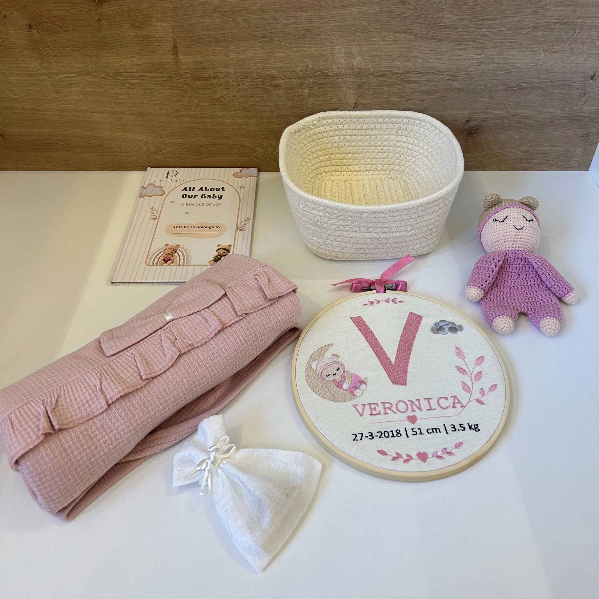 A baby shower gift set arranged on a white surface against a light wood background, including a pink textured blanket, a white drawstring bag, a woven basket, a personalized embroidery hoop with the name Veronica, a small plush doll, and a booklet.