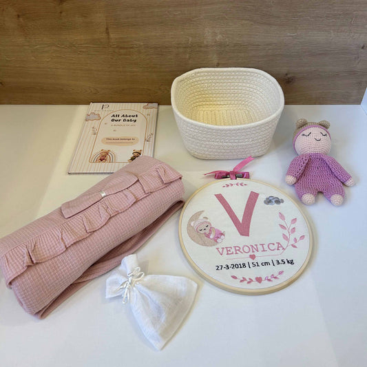 A baby shower gift set arranged on a white surface against a light wood background, including a pink textured blanket, a white drawstring bag, a woven basket, a personalized embroidery hoop with the name Veronica, a small plush doll, and a booklet.