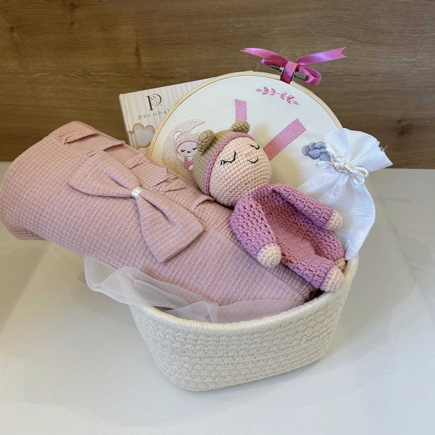 A baby gift basket containing a pink textured blanket with a large bow, a pink knitted bunny toy, a white embroidered hoop with a bunny design, a small white sachet, and a book, arranged in a woven basket against a wooden background.