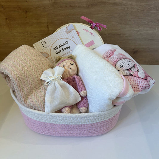 A baby gift basket containing a beige textured blanket, a white plush towel, a pink knitted baby pillow with a cartoon face, a small knitted doll, and a cream-colored drawstring pouch, all arranged in a pink and white woven basket against a wooden background.