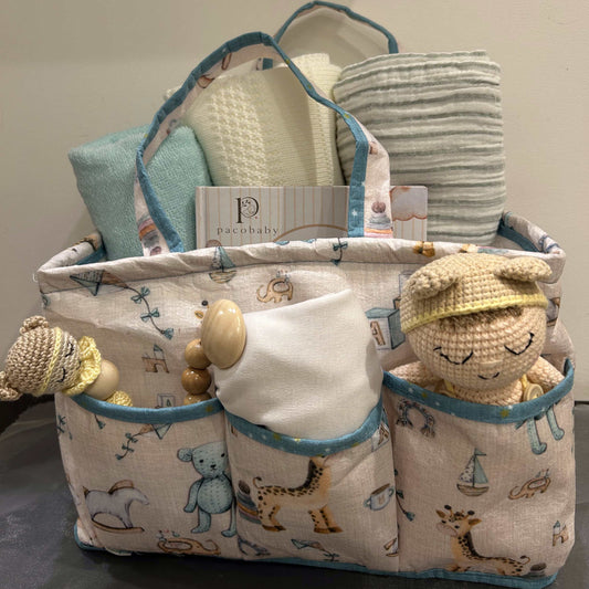 A fabric baby gift basket organizer with multiple pockets, featuring a cream and blue animal print, filled with baby items including a striped cloth, textured cream towel, and plush toys.