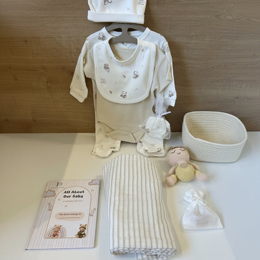A baby gift set arranged on a white surface against a wood-look background, including a cream and white long-sleeve bodysuit with a bib, a matching hat, folded striped blankets, a crochet doll, a small drawstring pouch, a woven basket, and a printed card reading 'All About Our Baby'.