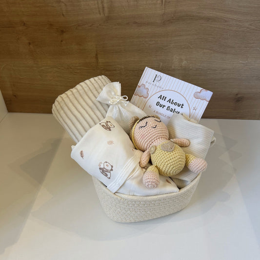 A baby gift basket set containing a cream-colored woven basket, a striped cream baby blanket, a white muslin swaddle with brown bunny prints, a small crocheted doll, and a book titled 'All About Our Baby'.