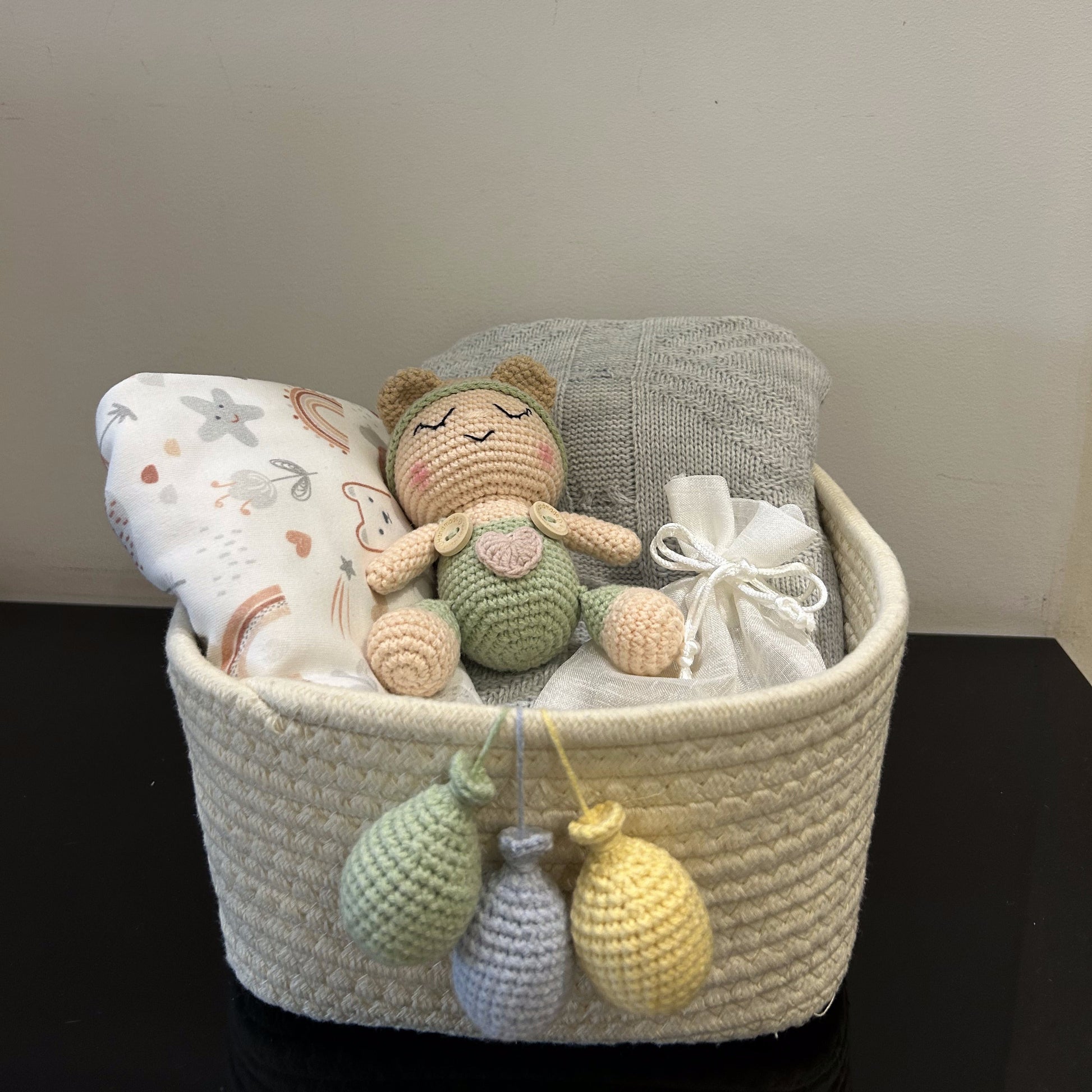 A woven cream baby gift basket filled with a crocheted doll, a patterned pillow, a gray cloth, and a white ribbon, sitting on a dark surface against a plain wall.