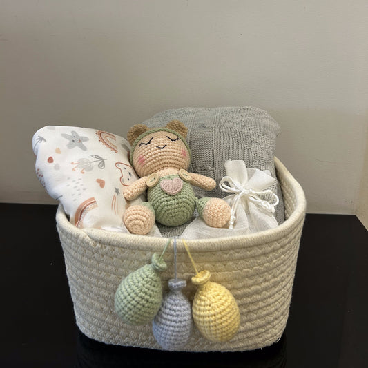 A woven cream baby gift basket filled with a crocheted doll, a patterned pillow, a gray cloth, and a white ribbon, sitting on a dark surface against a plain wall.
