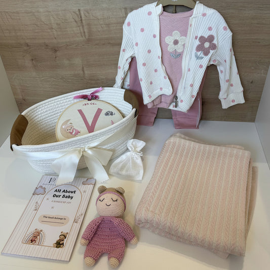 Baby gift set featuring a pink and white striped knit cardigan with floral appliques, a matching pink outfit, a white basket with a monogrammed baby hat, a folded cream knit blanket, a crocheted teddy bear, and a book.