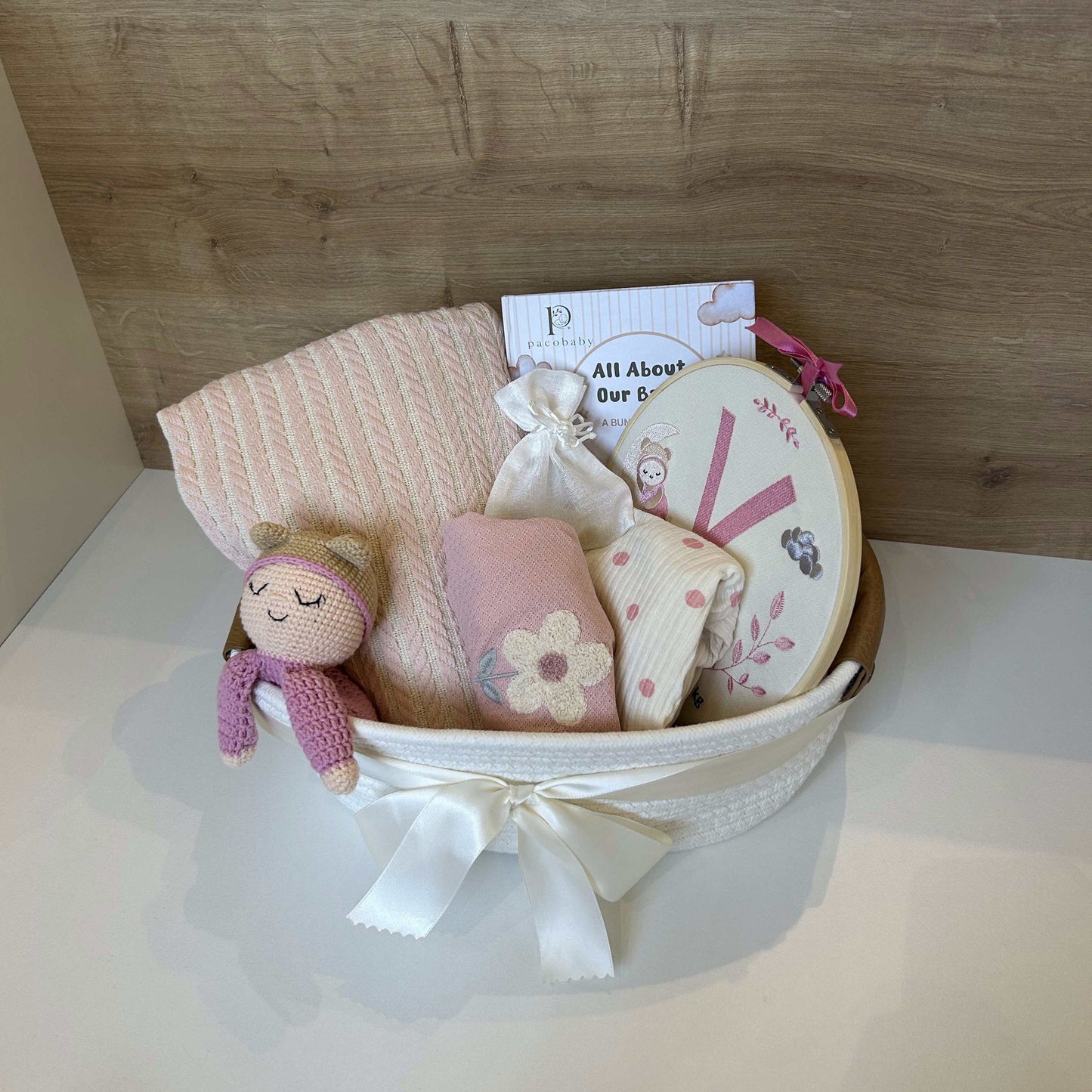 A baby gift basket containing a pink knit blanket, a small pink plush doll, folded baby clothes, a round mirror, and a card, tied with a white ribbon and set against a wood-paneled background.