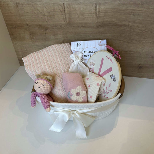 A baby gift basket containing a pink knit blanket, a small pink plush doll, folded baby clothes, a round mirror, and a card, tied with a white ribbon and set against a wood-paneled background.