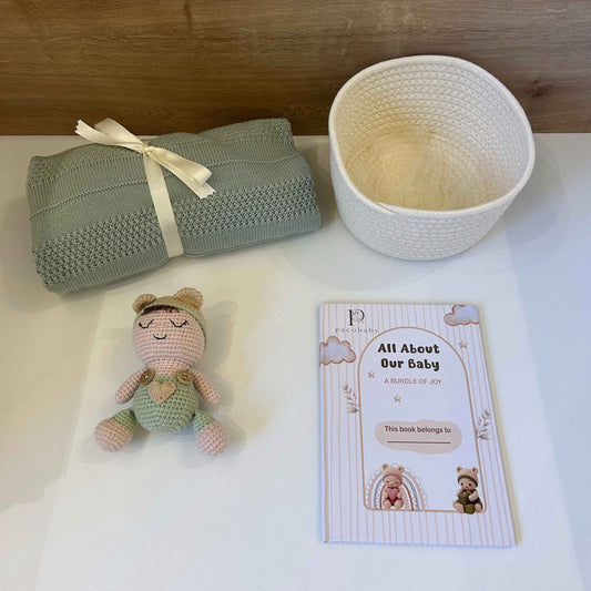 A baby gift set arranged on a white surface against a wood-grain background, including a folded sage green knit blanket tied with a cream ribbon, a cream woven basket, a small crocheted doll, and a printed card reading 'All About Our Baby'.