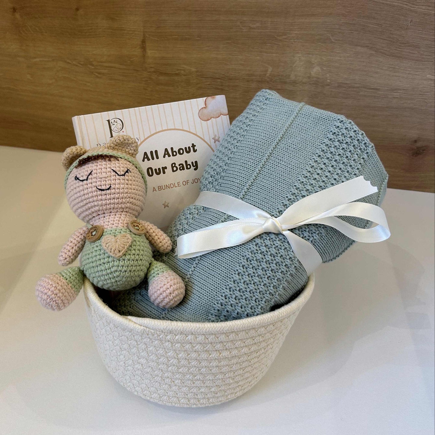 A baby gift set arranged in a white woven basket, featuring a light blue knitted blanket tied with a white ribbon, a small crocheted doll, and a card that reads 'All About Our Baby' against a wooden background.