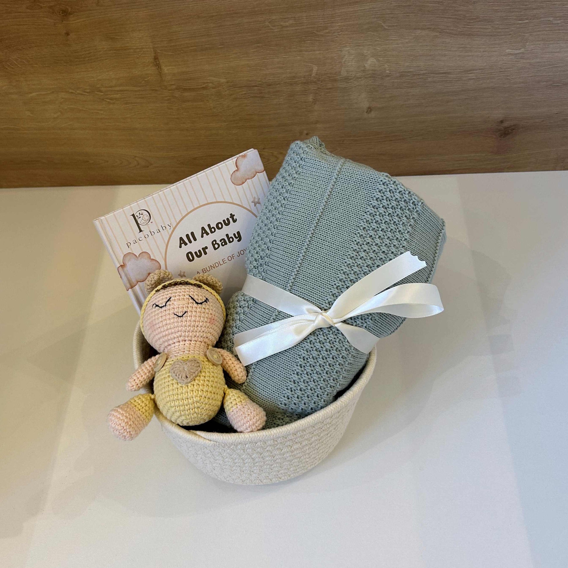 A baby gift set in a white textured bowl, featuring a folded sage green knit blanket tied with a white ribbon, a small crocheted baby doll, and a card that reads 'All About Our Baby'.
