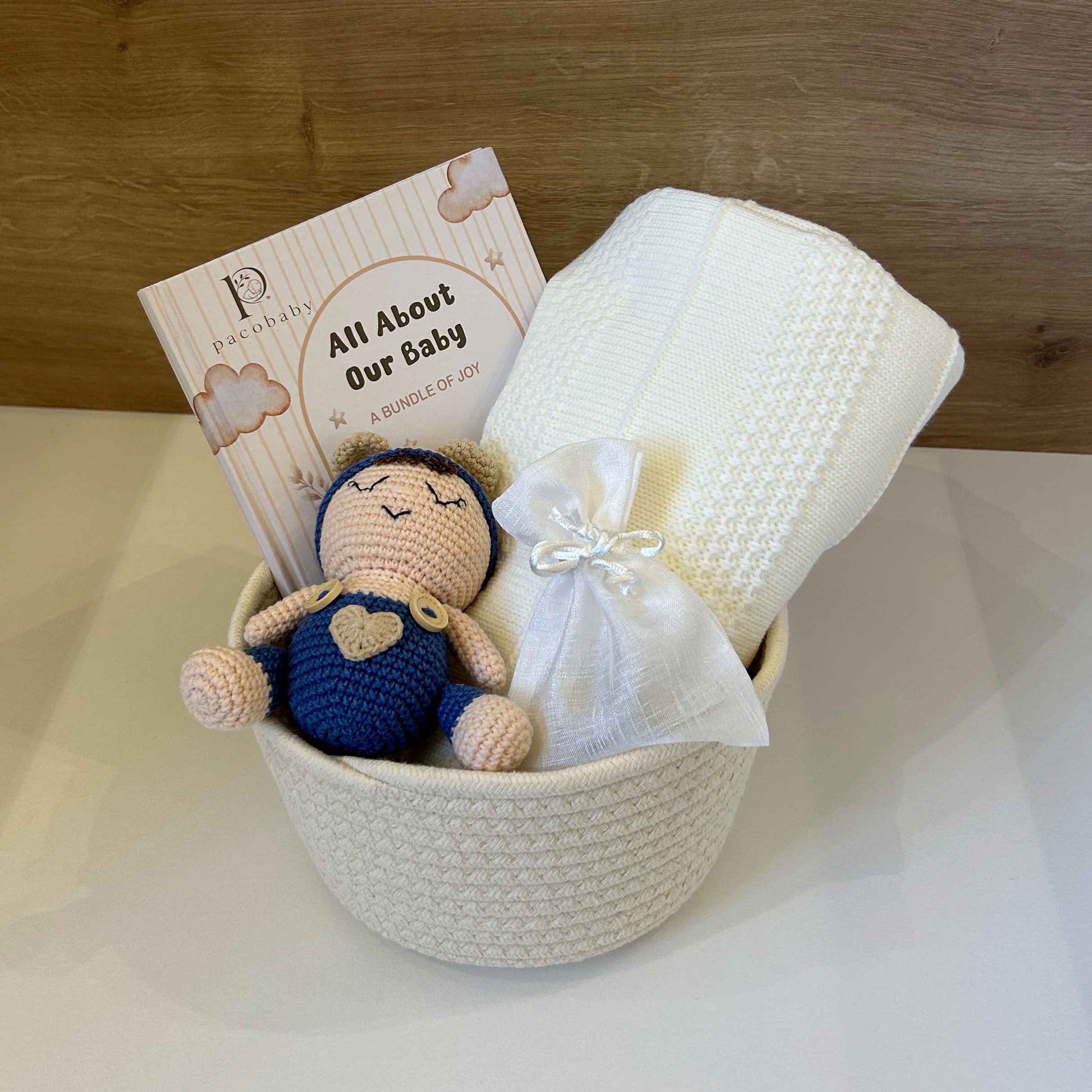 A baby gift basket set in a white woven bowl, featuring a crocheted baby doll, a white textured blanket, a white swaddle wrap, and a printed card that reads 'All About Our Baby'. The basket is set against a light-colored surface and a wooden wall background.