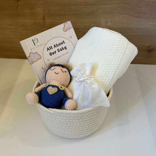 A baby gift basket set in a white woven bowl, featuring a crocheted baby doll, a white textured blanket, a white swaddle wrap, and a printed card that reads 'All About Our Baby'. The basket is set against a light-colored surface and a wooden wall background.