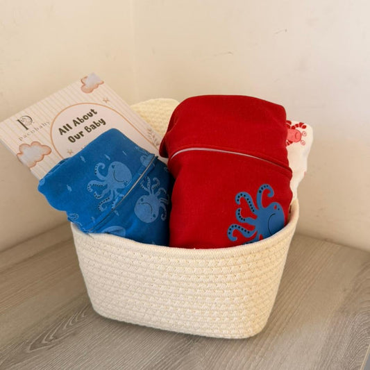 A baby gift basket containing folded red and blue clothing with octopus embroidery, a blue cloth, and a card that reads 'All Aboard Our Baby', placed in a white woven basket on a light wood surface against a beige wall.