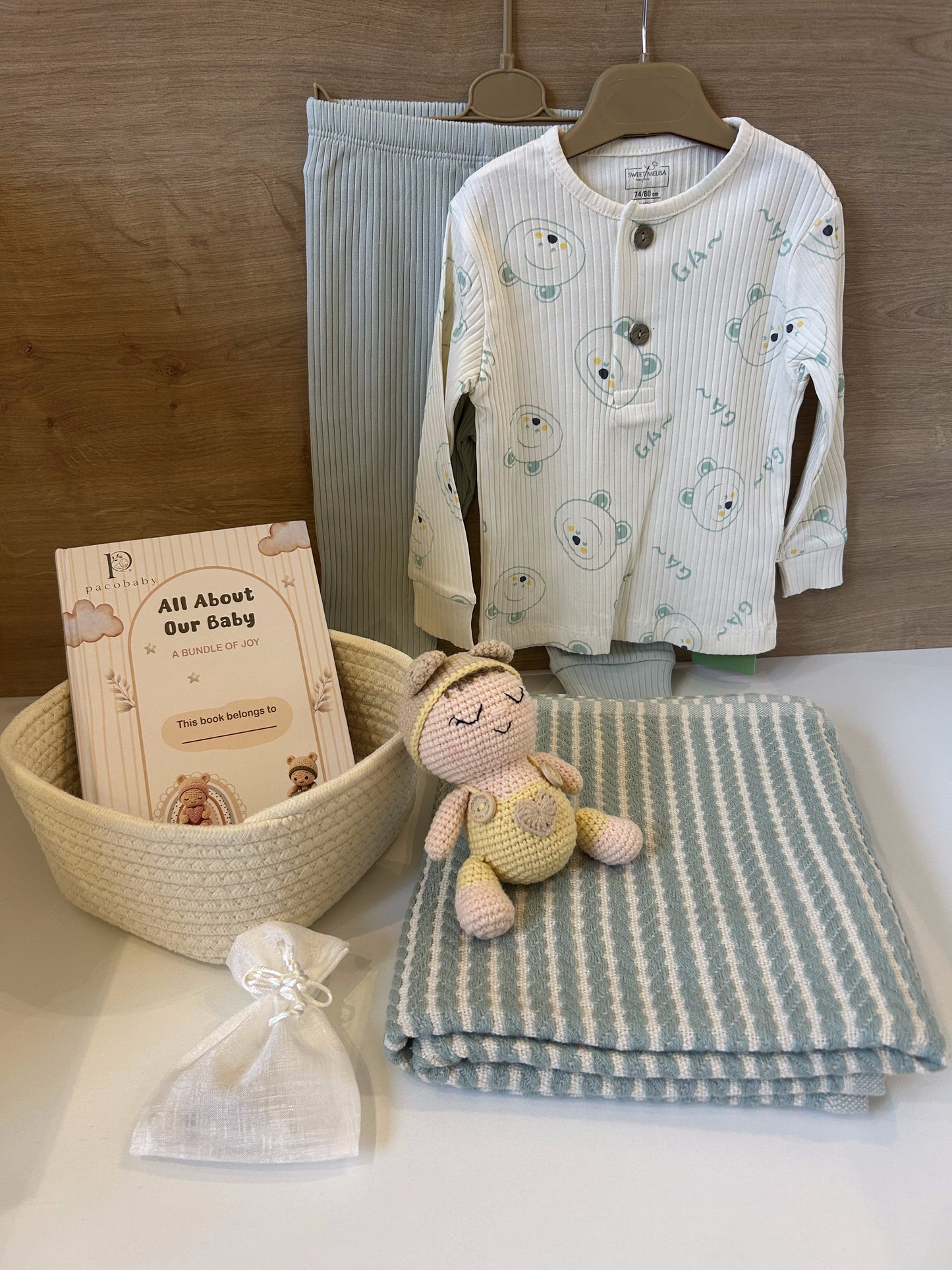 A baby gift set arranged on a white surface against a wood wall, including a cream ribbed long-sleeve top with bear face prints, a pair of light blue striped pants, a folded blue-and-cream striped blanket, a small crocheted doll, a woven basket with a book, and a small white drawstring pouch.