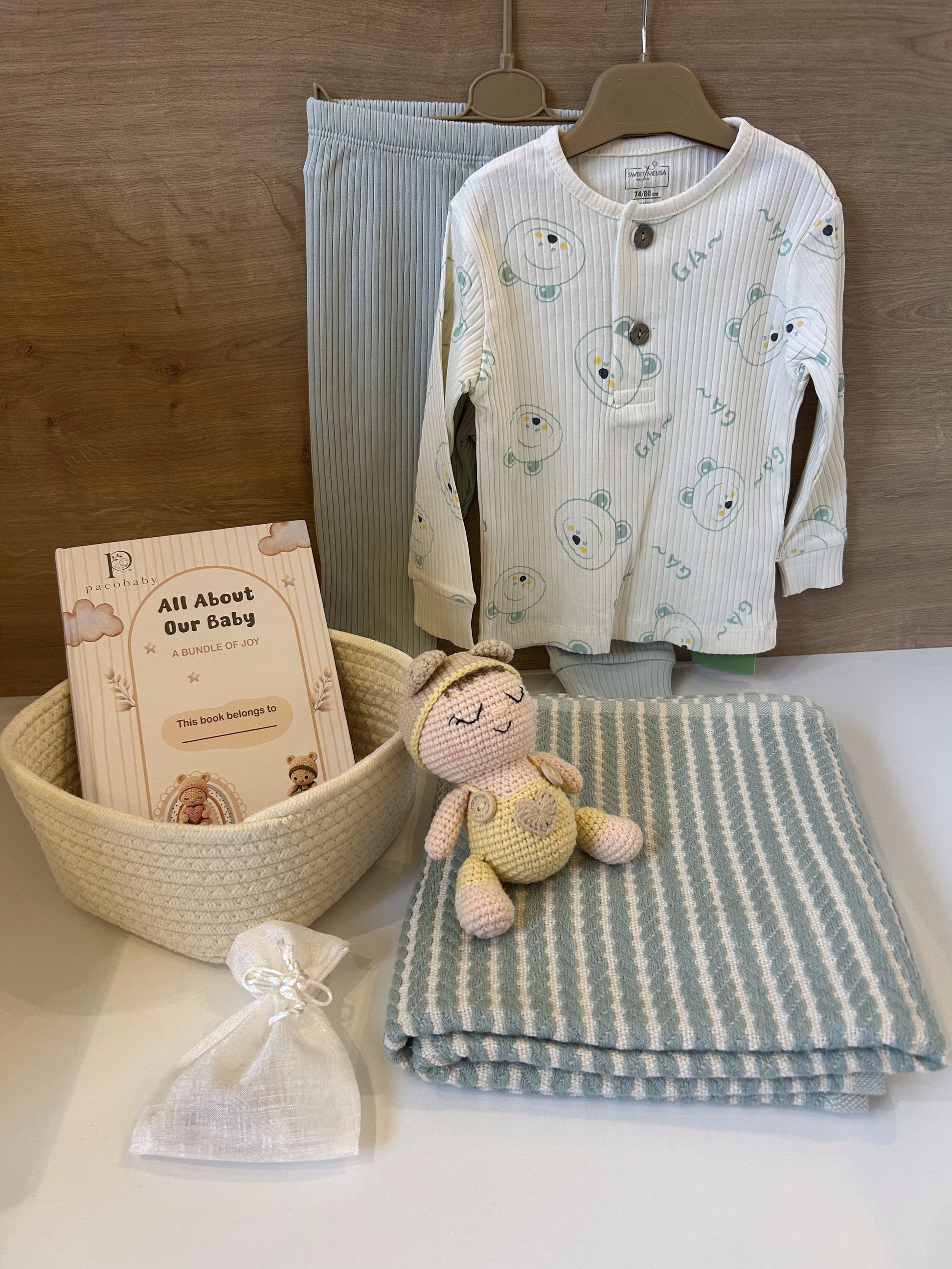A baby gift set arranged on a white surface against a wood wall, including a cream ribbed long-sleeve top with bear face prints, a pair of light blue striped pants, a folded blue-and-cream striped blanket, a small crocheted doll, a woven basket with a book, and a small white drawstring pouch.