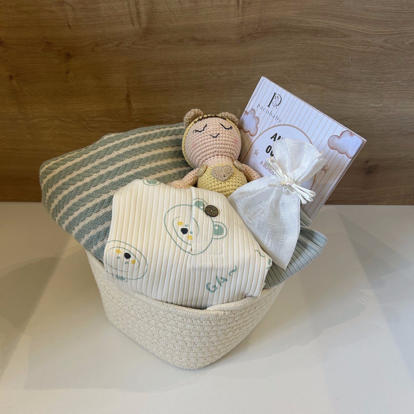 A baby gift basket set in a woven cream basket, containing a striped green and white blanket, a cream muslin swaddle with embroidered cartoon faces, a small crocheted teddy bear toy, and a small white drawstring pouch with a card, set against a wooden wall background.
