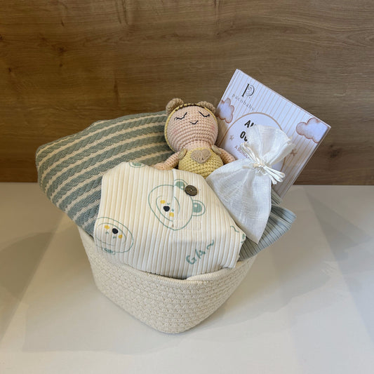 A baby gift basket set in a woven cream basket, containing a striped green and white blanket, a cream muslin swaddle with embroidered cartoon faces, a small crocheted teddy bear toy, and a small white drawstring pouch with a card, set against a wooden wall background.