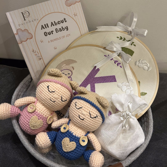 A baby keepsake gift set in a gray felt basket, featuring two crocheted baby dolls, embroidered round photo frames, a white fabric drawstring pouch, and a printed booklet titled 'All About Our Baby' by Pashmina.