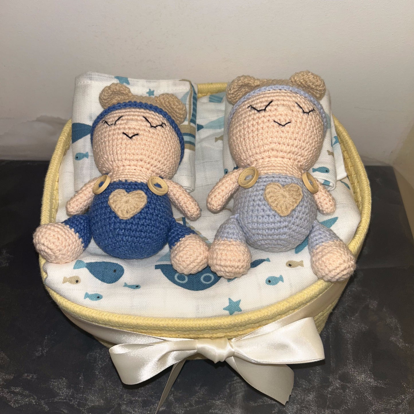 Two crocheted baby doll toys sitting side by side in a round fabric basket with a white satin bow. The doll on the left wears a blue outfit with a beige heart, and the doll on the right wears a light blue outfit with a beige heart.