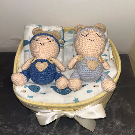 Two crocheted baby doll toys sitting side by side in a round fabric basket with a white satin bow. The doll on the left wears a blue outfit with a beige heart, and the doll on the right wears a light blue outfit with a beige heart.