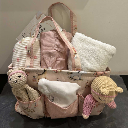 A pink fabric diaper bag with multiple front pockets, filled with baby essentials including a white towel, pink cloth items, and a white cloth napkin, with two small crocheted bunny toys hanging from the sides.