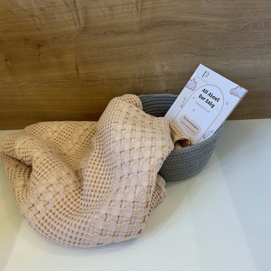 A beige woven baby blanket draped over a gray basket, with a printed insert card partially visible inside.