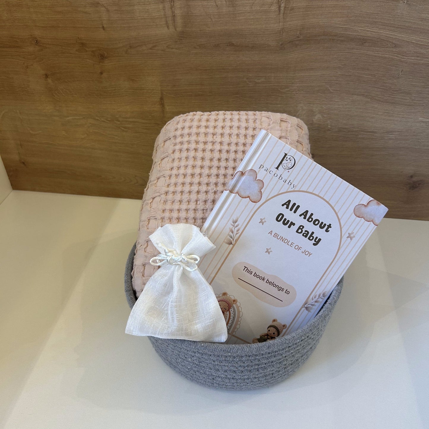 A baby keepsake gift set in a gray crocheted basket, featuring a pink and white checkered blanket, a small white fabric pouch tied with a bow, and a printed card that reads 'All About Our Baby' by P.ashley.