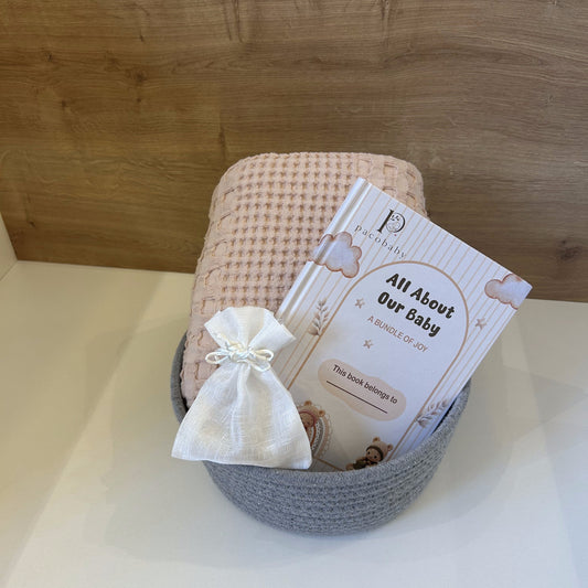 A baby keepsake gift set in a gray crocheted basket, featuring a pink and white checkered blanket, a small white fabric pouch tied with a bow, and a printed card that reads 'All About Our Baby' by P.ashley.