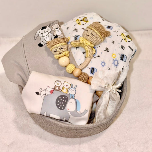 A baby gift set arranged in a round gray basket, including a knitted yellow and beige baby teething ring with wooden beads, a white muslin blanket with colorful cartoon icons, a gray cloth with a cartoon animal print, and a white fabric pouch tied with a ribbon.