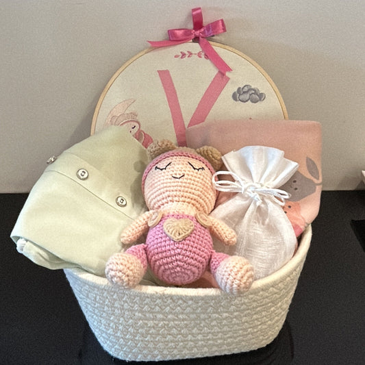 A baby gift basket arrangement featuring a crocheted pink bunny toy, a cream-colored knit hat, a folded cream garment, a white drawstring bag, and a round decorative plate with a pink ribbon bow, set against a dark background.