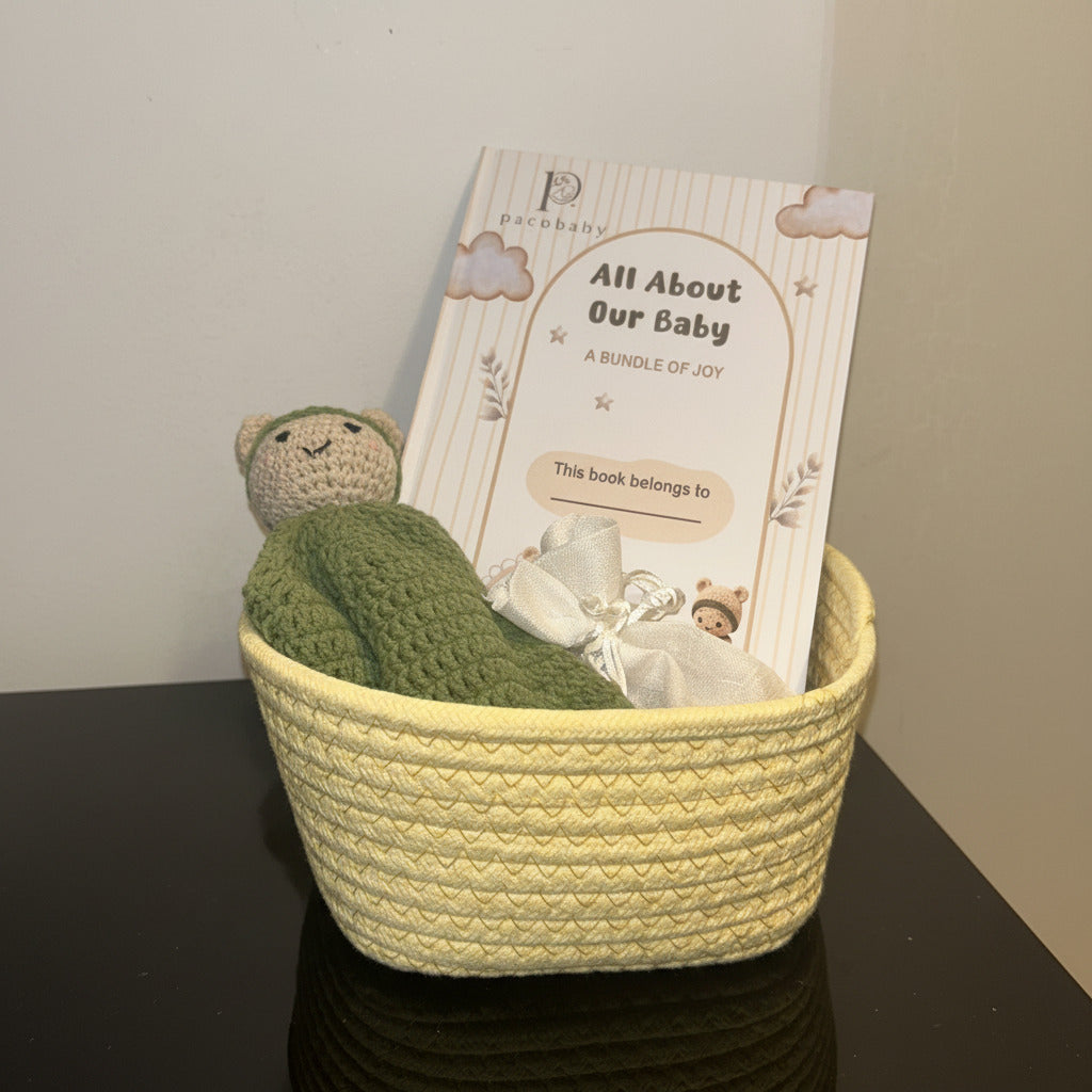 A baby book titled 'All About Our Baby' resting inside a yellow woven basket, decorated with a green crocheted hat and a small plush toy on a plain white background.