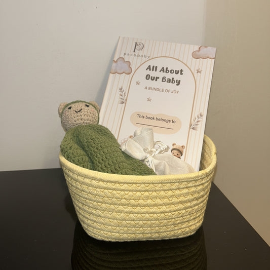 A baby book titled 'All About Our Baby' resting inside a yellow woven basket, decorated with a green crocheted hat and a small plush toy on a plain white background.