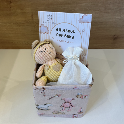 A baby keepsake gift basket set from Pearlylakes, featuring a small patterned fabric box with a crocheted baby doll, a white drawstring pouch, and a printed card that reads 'All About Our Baby'.