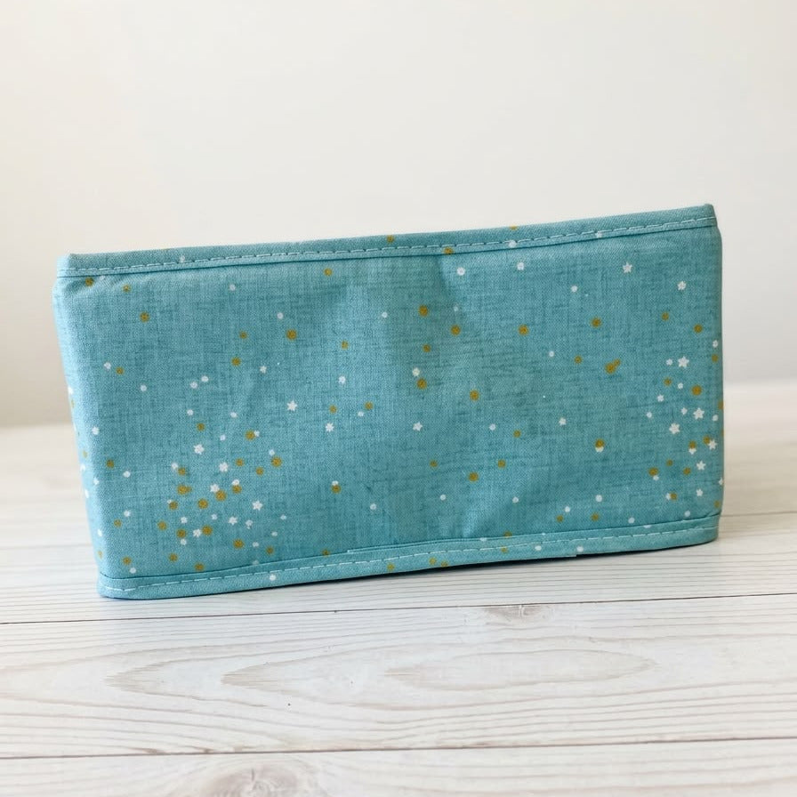 A teal fabric pouch with a gold and white speckled star pattern, resting on a light-colored wooden surface against a neutral background.