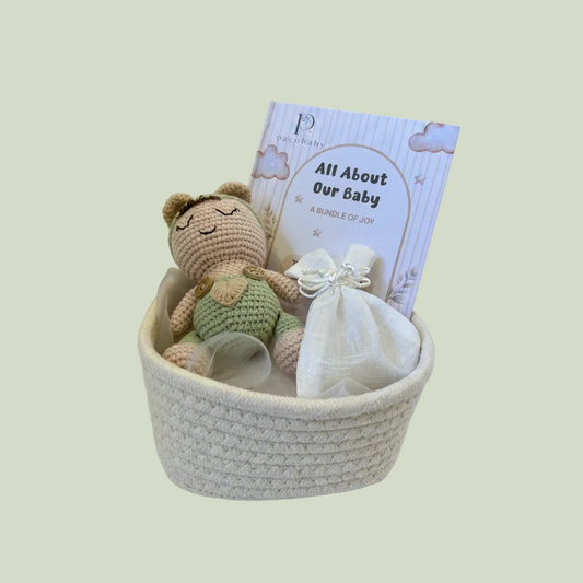 A baby gift basket set containing a crocheted teddy bear doll, a small white fabric pouch tied with a ribbon, and a printed card that reads 'All About Our Baby' with the brand name 'pachababy' visible, all arranged inside a cream woven basket.