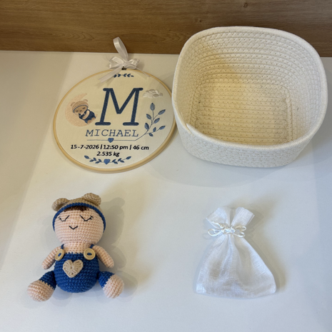 A baby keepsake gift set featuring a crocheted doll in blue and beige, a small white fabric pouch tied with a bow, a cream woven basket, and a personalized round wooden ornament with the name 'MICHAEL' and monogram 'M'.