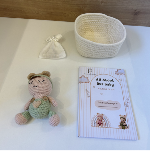 A baby keepsake gift set featuring a crocheted doll, a white woven basket, a small cream drawstring pouch, and a personalized 'All About Our Baby' photo book, arranged on a white surface against a wooden background.