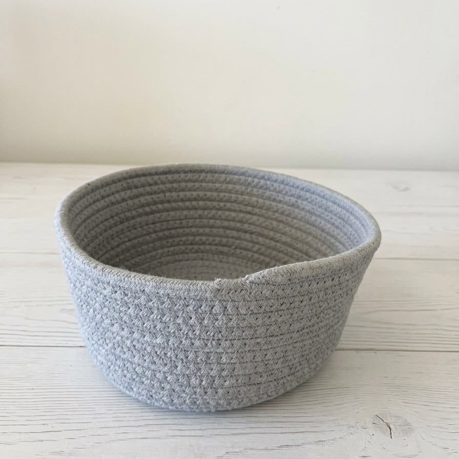 A light gray crocheted storage basket with a ribbed texture, sitting on a white wooden surface against a plain off-white wall.
