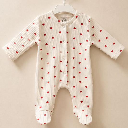 A white ribbed knit baby footed sleeper with a red heart pattern, hanging on a white plastic hanger against a solid beige background.
