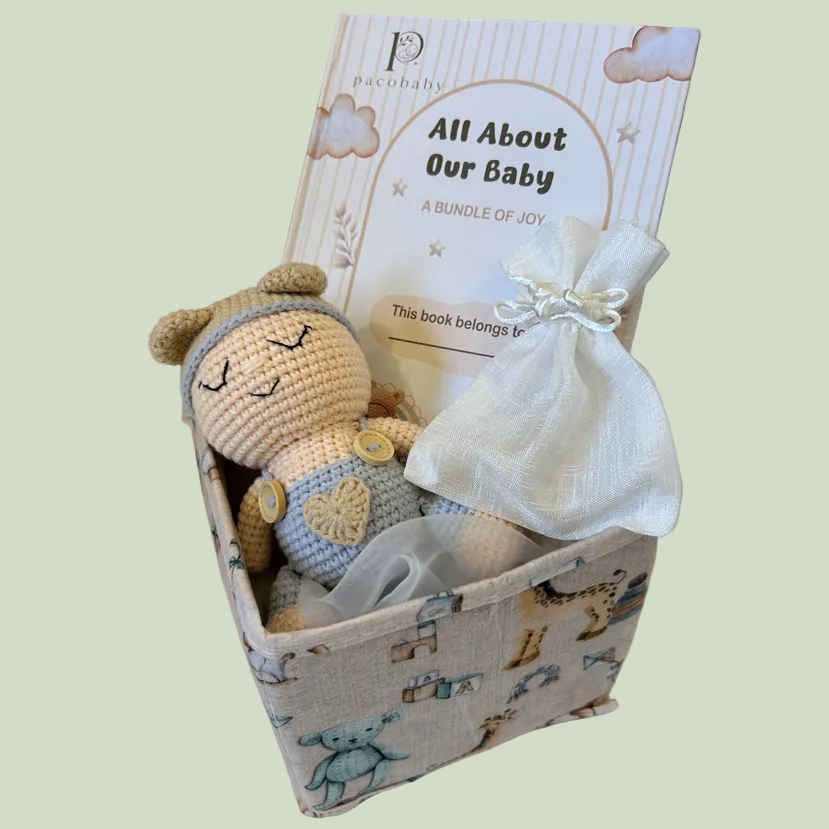 A baby gift set in a patterned fabric box featuring a crocheted doll, a white drawstring pouch, and a printed booklet titled 'All About Our Baby'.